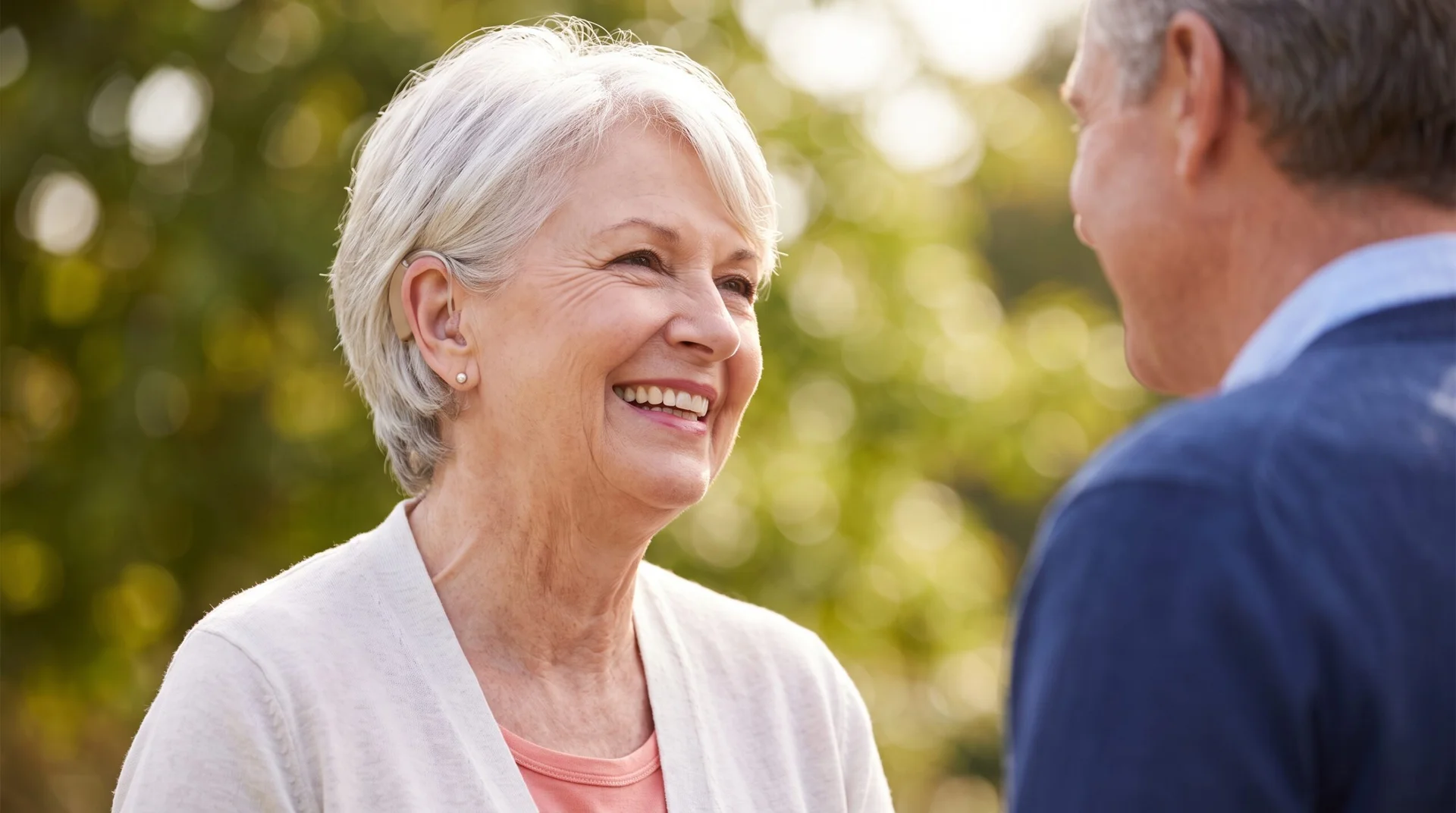 A smiling senior woman wearing an advanced hearing solution enjoys a conversation outdoors in natural light.